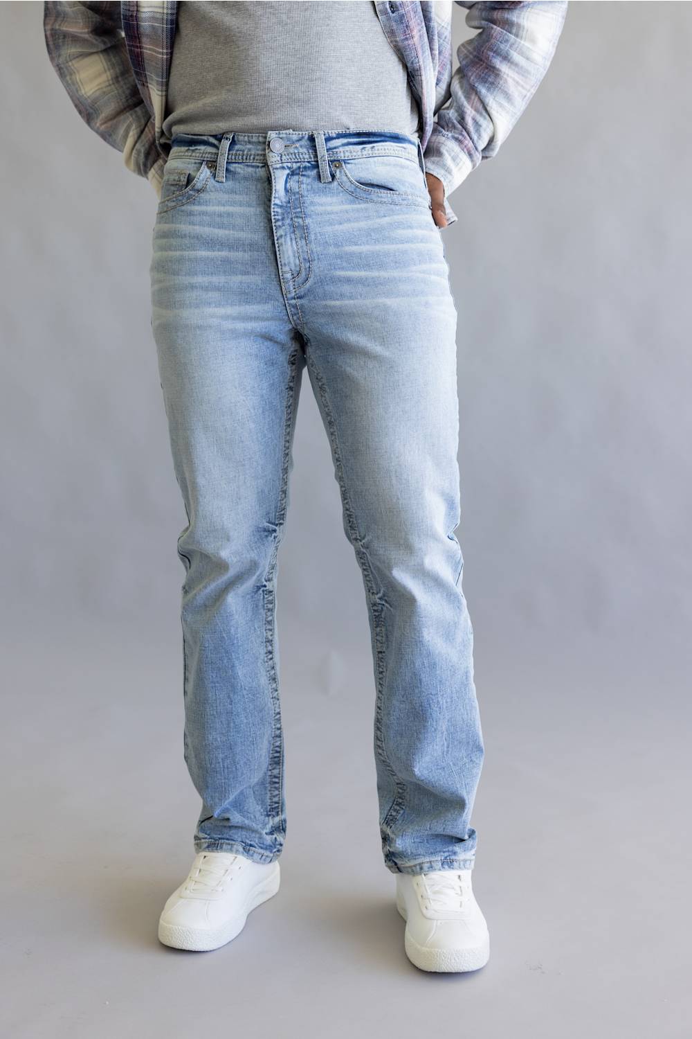 A person wearing light-wash blue jeans, a gray t-shirt, a plaid flannel shirt, and white sneakers, standing against a neutral gray studio background.