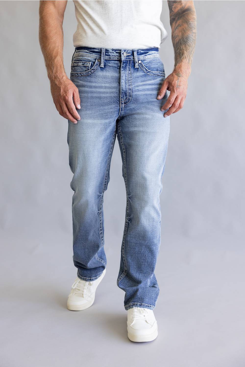 Rear view of a person wearing light blue denim jeans with decorative embroidered patch detailing on the back pockets, paired with a white t-shirt against a neutral gray background.