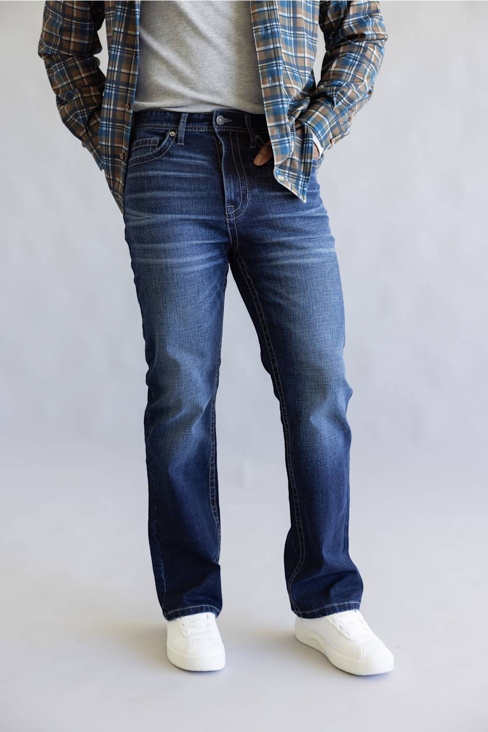 A person wearing dark blue straight-leg denim jeans, styled with a gray t-shirt, a blue and brown plaid flannel shirt, and white sneakers, standing against a light gray studio background.