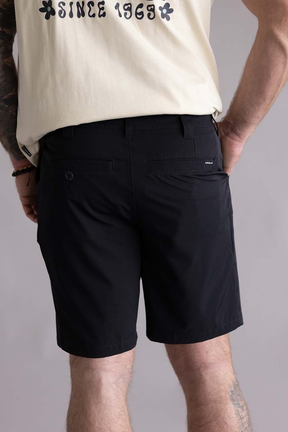 Back view of a person wearing black shorts with two visible pockets and a beige shirt with text on a gray background. The shorts are the Quiksilver Taxer Amphibian 18" Hybrid Shorts for Men in Black 