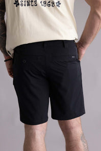 Back view of a person wearing black shorts with two visible pockets and a beige shirt with text on a gray background. The shorts are the Quiksilver Taxer Amphibian 18" Hybrid Shorts for Men in Black 