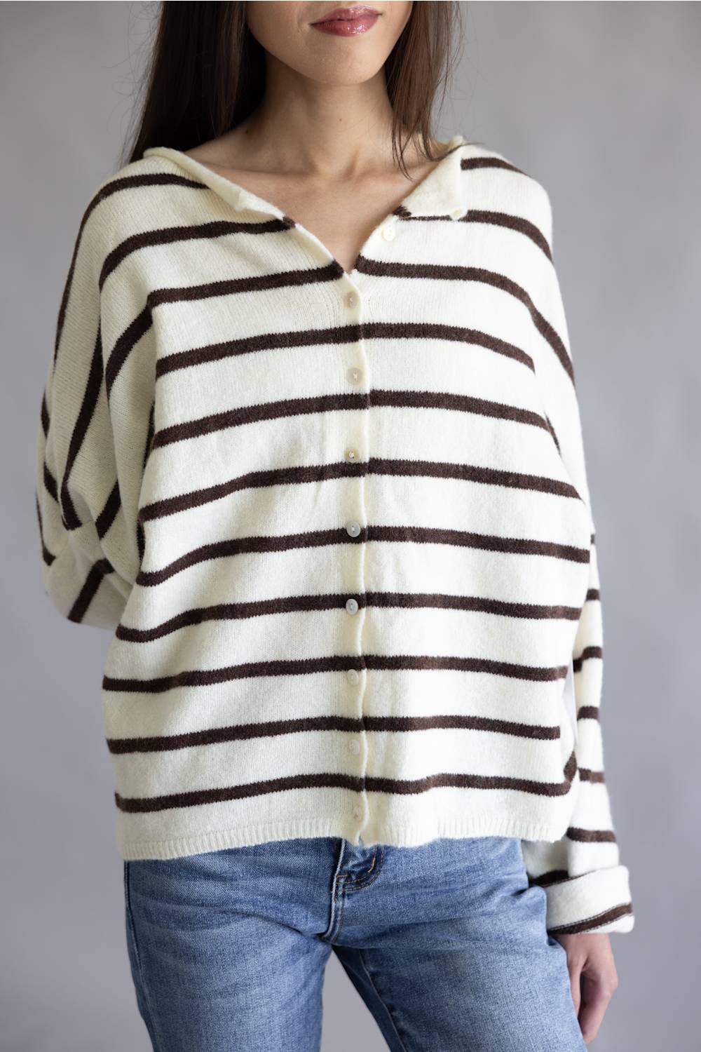 A woman wearing a cream knit cardigan with dark brown horizontal stripes, paired with blue jeans, shown against a plain gray studio background.