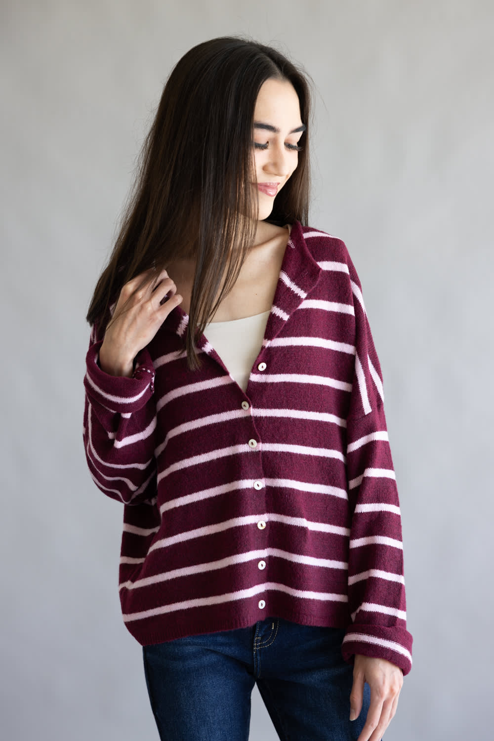 A woman wearing a burgundy and light pink striped button-up cardigan over a cream top and dark blue jeans, standing against a neutral gray studio background.