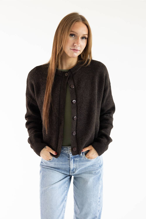 A woman with long light brown hair stands against a plain white studio background wearing a dark brown button-up cardigan over an olive green top and light blue jeans.
