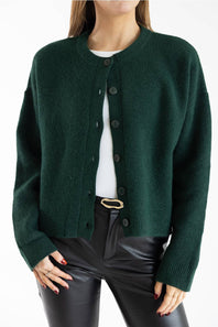A woman wearing a dark forest green knit cardigan with a crew neck and dark buttons, layered over a white top and paired with black pants.