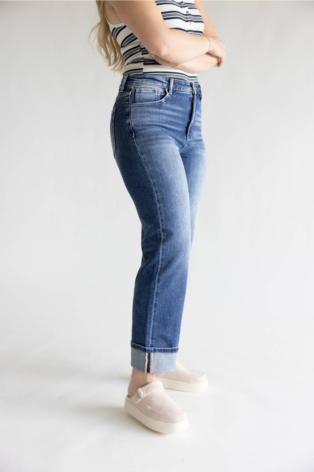 A person wearing medium-wash blue cuffed jeans, a striped tank top, and cream slip-on shoes, standing against a plain light gray studio background.