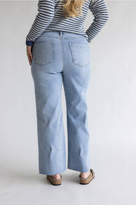 A person shown from the back wearing light-wash blue wide-leg jeans, a navy and white striped long-sleeve top, and brown flat shoes against a plain light gray studio background.