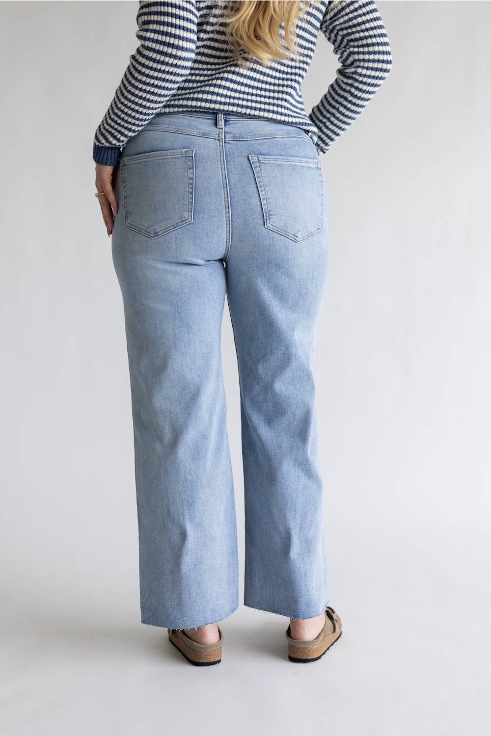 A person shown from the back wearing light-wash blue wide-leg jeans, a navy and white striped long-sleeve top, and brown flat shoes against a plain light gray studio background.