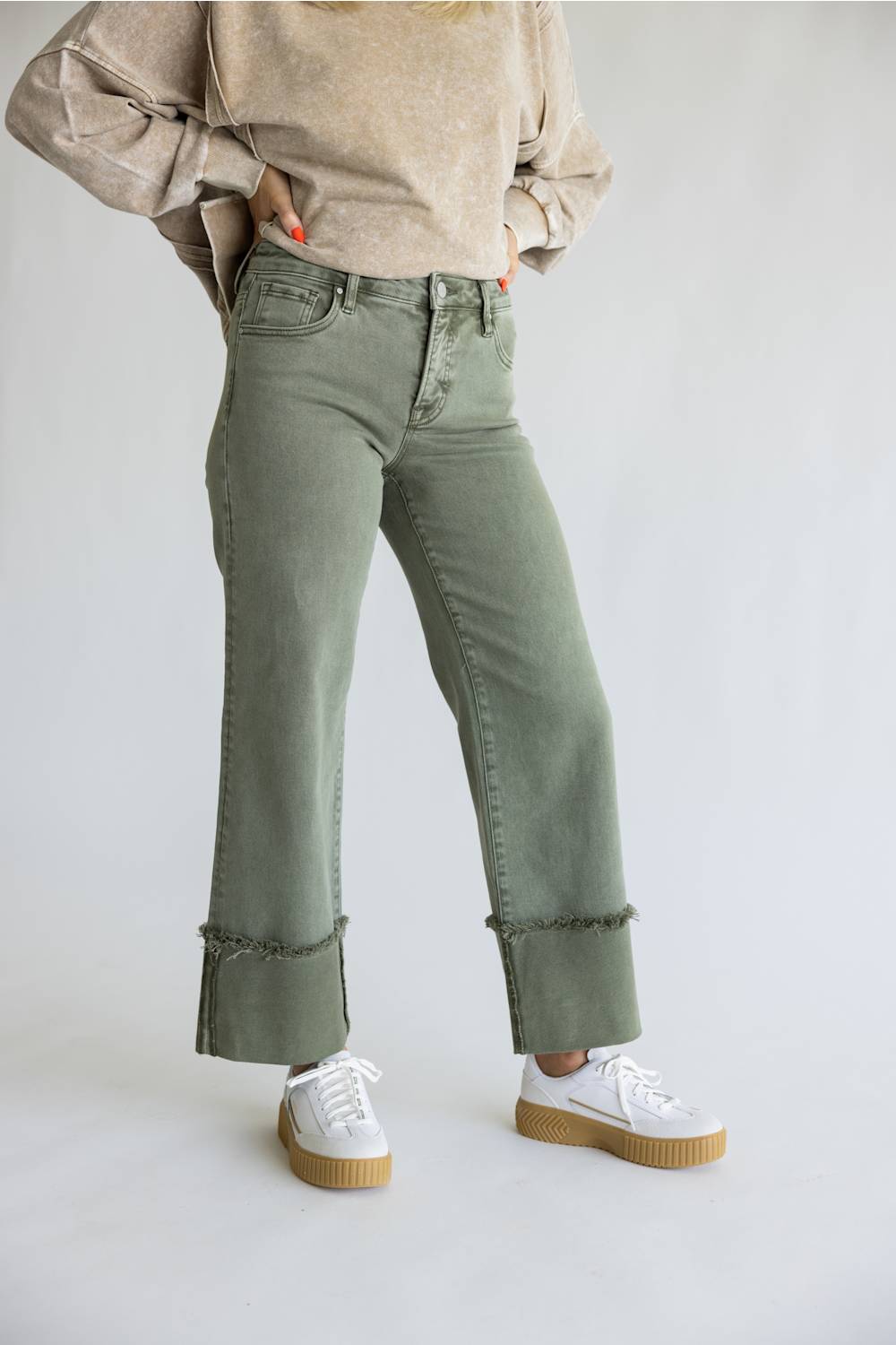 A person wearing sage green wide-leg cropped jeans with rolled cuffs, paired with a beige sweater and white platform sneakers, standing against a light gray studio background.