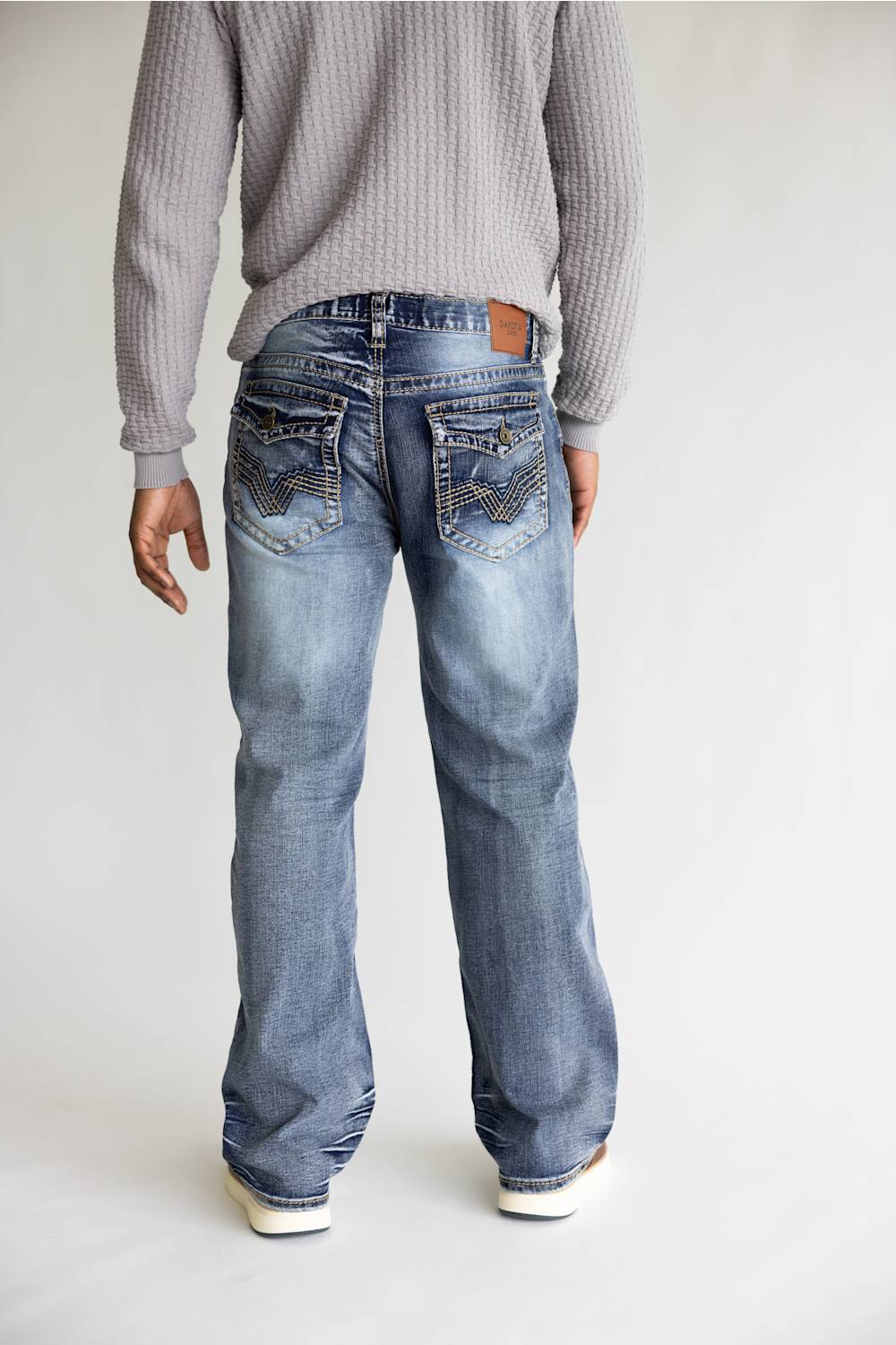 A person shown from the back wearing light-wash blue jeans with decorative stitching on the back pockets and a gray knit sweater, standing against a plain light background.