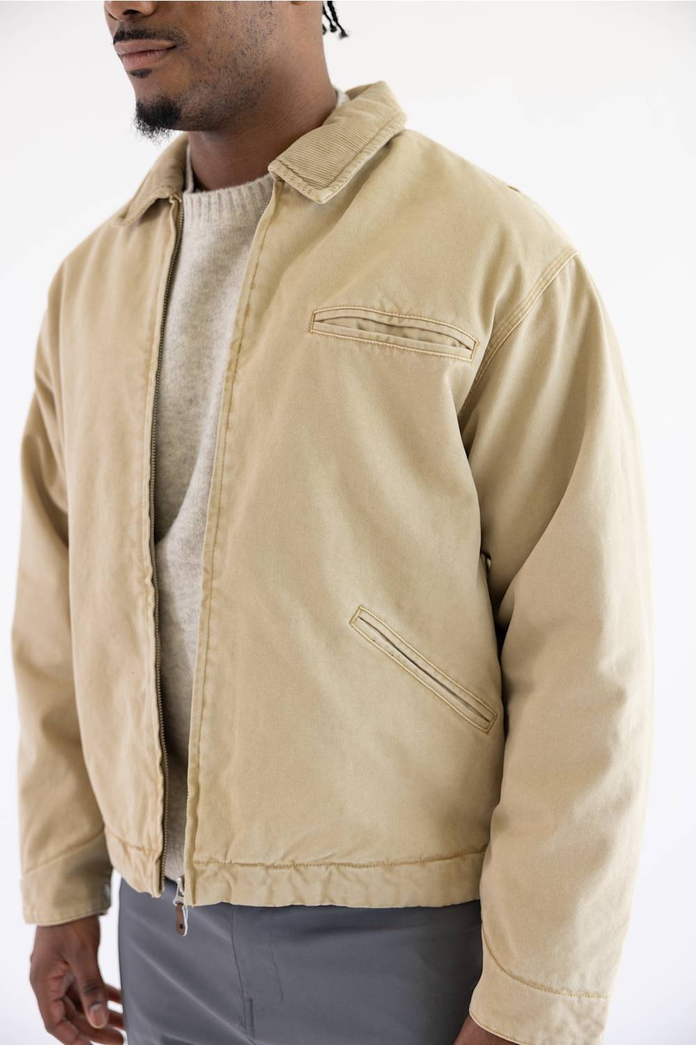 A man wearing a light beige canvas utility jacket over a light gray sweater and gray pants, shown from the chin down against a plain white background.