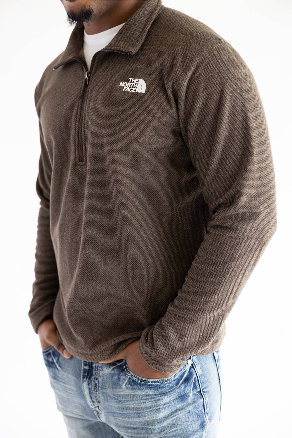 A man wearing a brown quarter-zip fleece pullover by The North Face, paired with blue jeans, shown from the chest down against a white background.