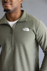 A close-up of a man wearing a heathered olive green half-zip pullover by The North Face, layered over a white t-shirt, against a neutral gray background.