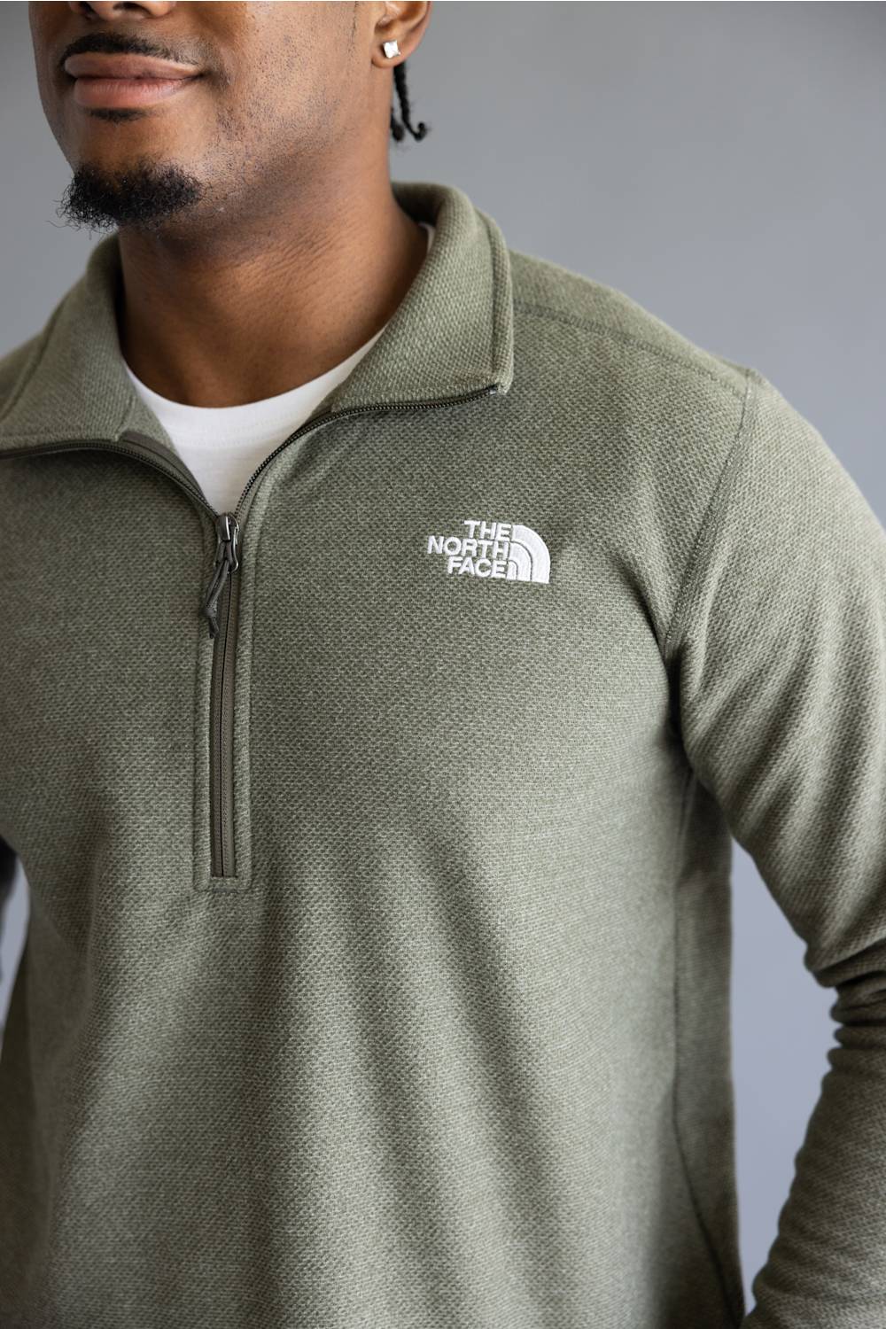 A close-up of a man wearing a heathered olive green half-zip pullover by The North Face, layered over a white t-shirt, against a neutral gray background.
