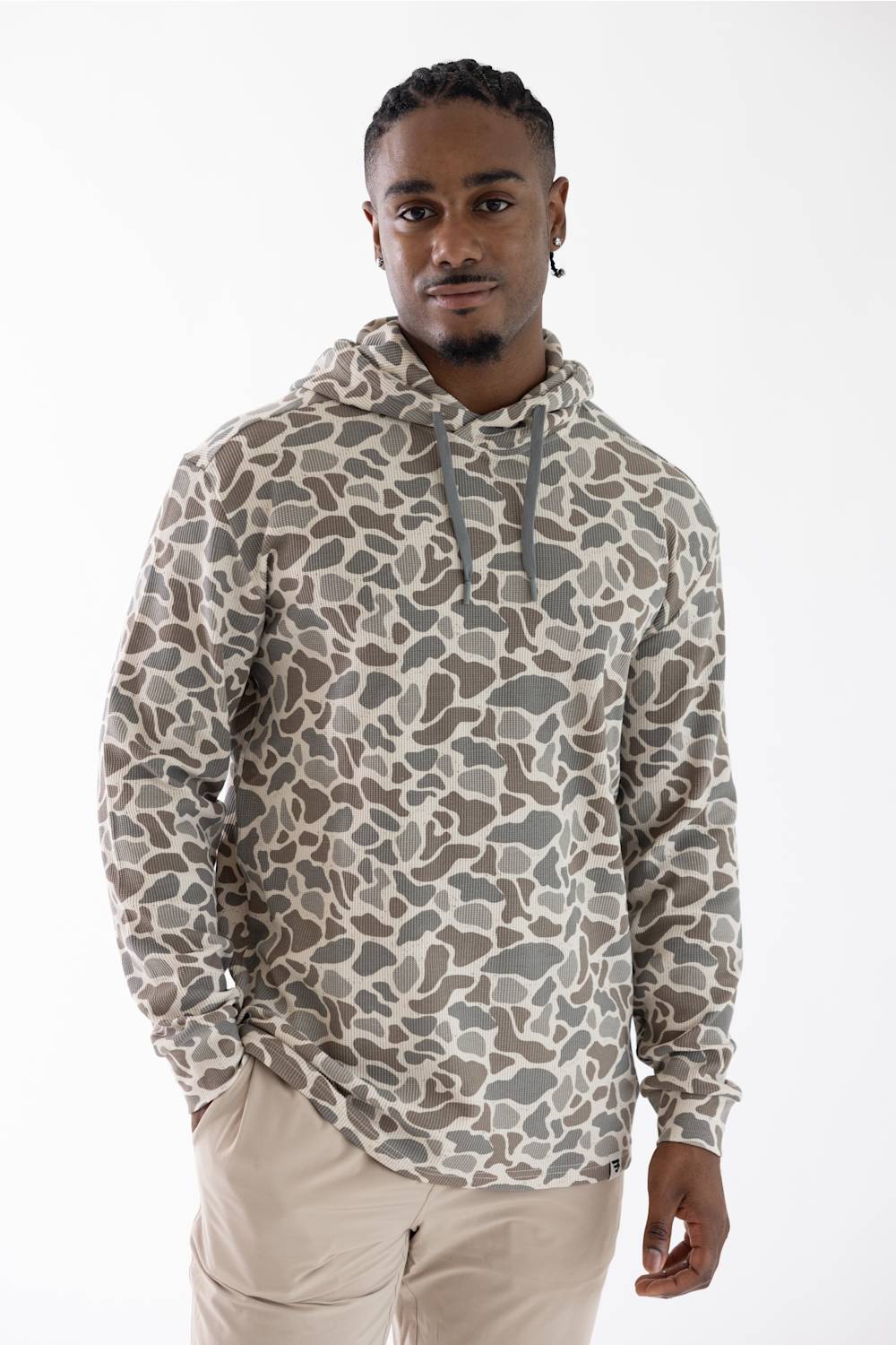A man wearing a beige and gray camouflage patterned hoodie with drawstrings, paired with beige pants, standing against a plain white studio background.