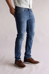 Side view of a person wearing blue jeans and brown shoes on a plain background. The jeans are Weatherproof Vintage Slim Jeans for Men in Tidepool Tint.