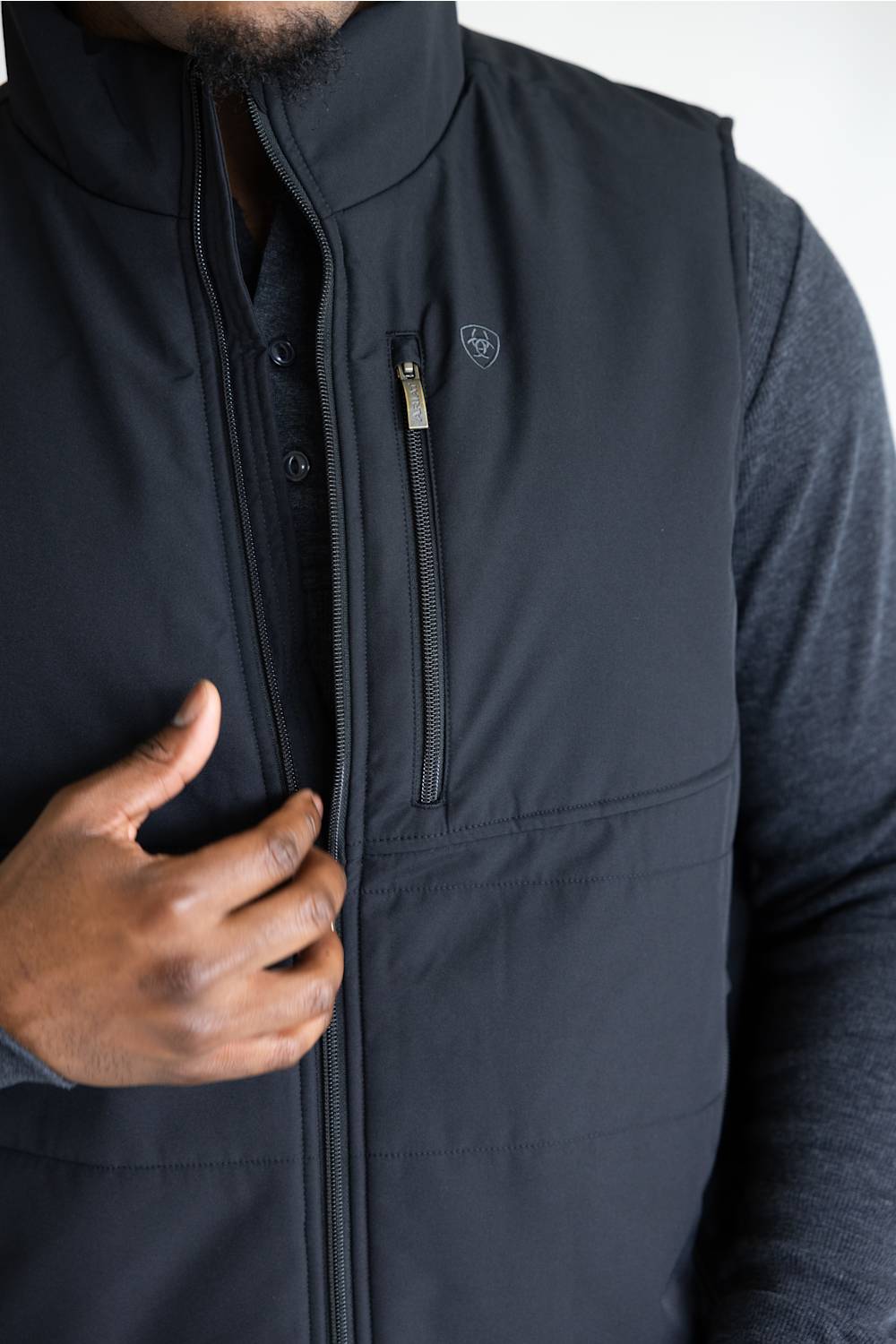 Close-up of a person wearing a black zip-front vest over a dark long-sleeve shirt, with one hand pulling the zipper. A small shield-style logo is visible on the chest pocket.