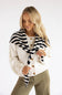 A blonde woman wearing a cream knit cardigan with brown buttons, a black and white striped knit scarf, and olive green pants, photographed against a clean white studio background.