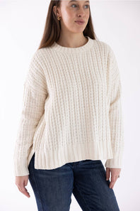 A woman wearing a cream-colored, long-sleeve ribbed knit sweater and dark blue jeans, photographed against a plain white studio background.