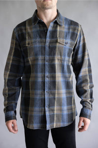 A male model wearing the Split Flannel Shirt for Men in Moka, featuring a blue, dark gray, and tan plaid pattern with long sleeves, dual buttoned chest pockets, and a button-down front, paired with black pants against a light gray studio background.