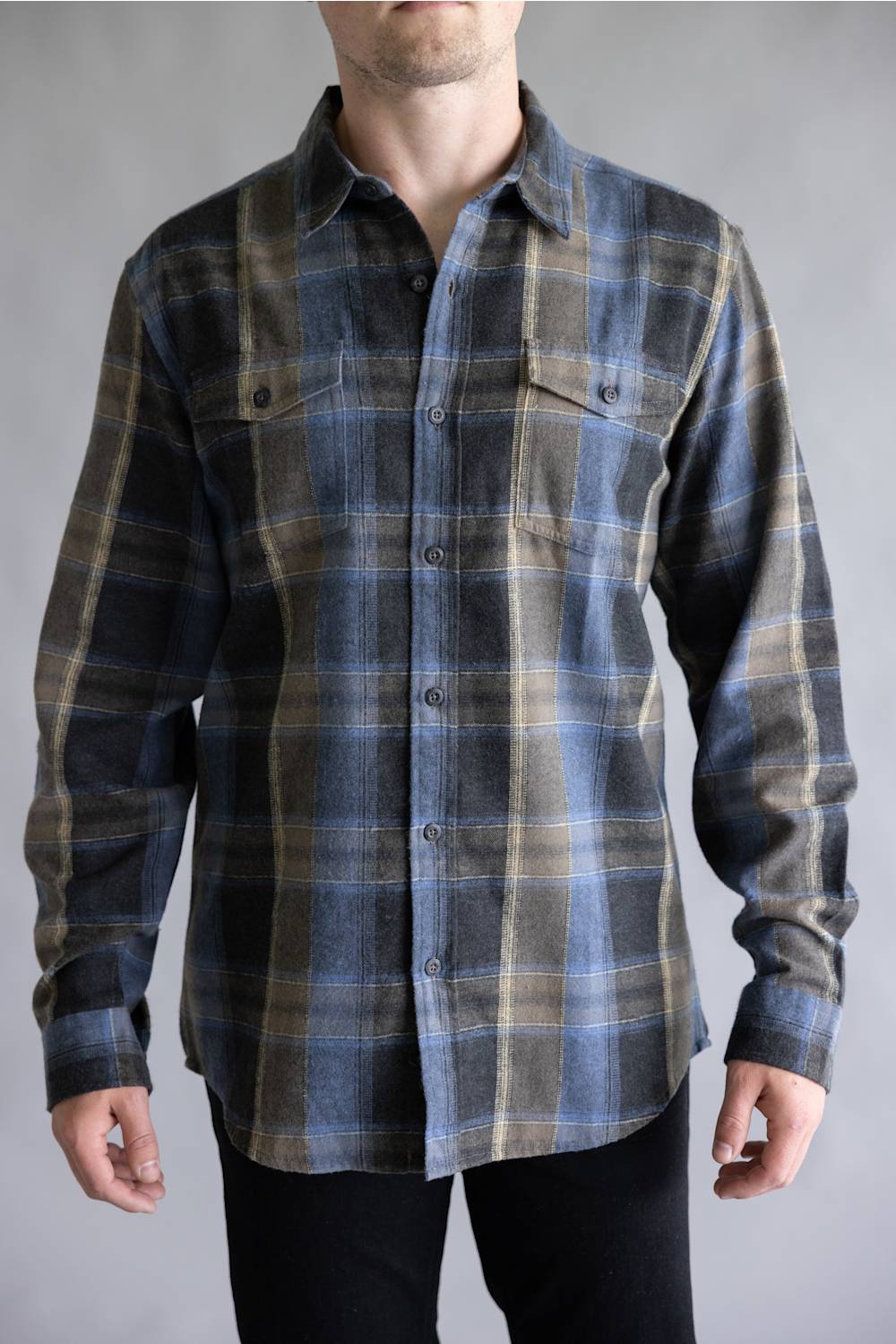 A male model wearing the Split Flannel Shirt for Men in Moka, featuring a blue, dark gray, and tan plaid pattern with long sleeves, dual buttoned chest pockets, and a button-down front, paired with black pants against a light gray studio background.