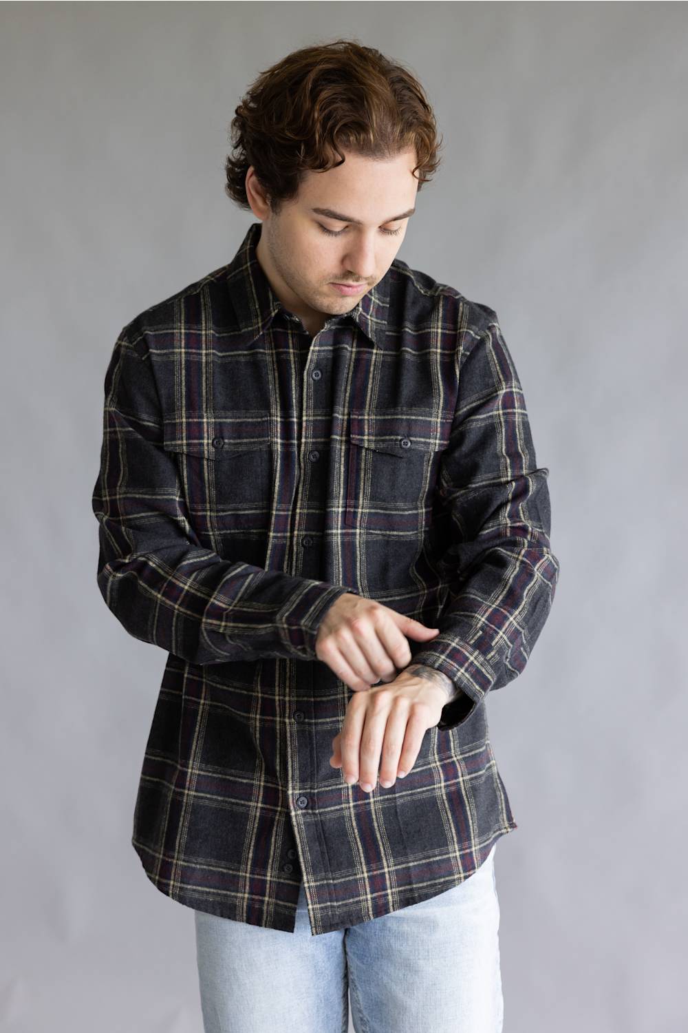 A male model adjusting the cuff of his sleeve while wearing the Split Flannel Shirt for Men in Slate, featuring a charcoal gray plaid pattern with burgundy and cream accent lines, long sleeves, and dual chest button pockets, paired with light wash jeans against a plain gray studio background.