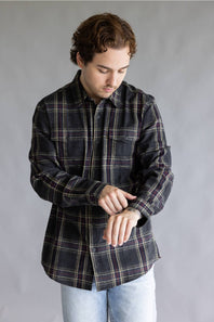 A male model adjusting the cuff of his sleeve while wearing the Split Flannel Shirt for Men in Slate, featuring a charcoal gray plaid pattern with burgundy and cream accent lines, long sleeves, and dual chest button pockets, paired with light wash jeans against a plain gray studio background.