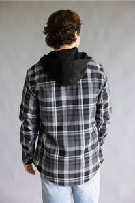 Back view of a model wearing the Split Men's long-sleeve hooded flannel shirt in a black, grey, and white plaid pattern, featuring a solid black attached hood and a straight back yoke against a grey studio background.