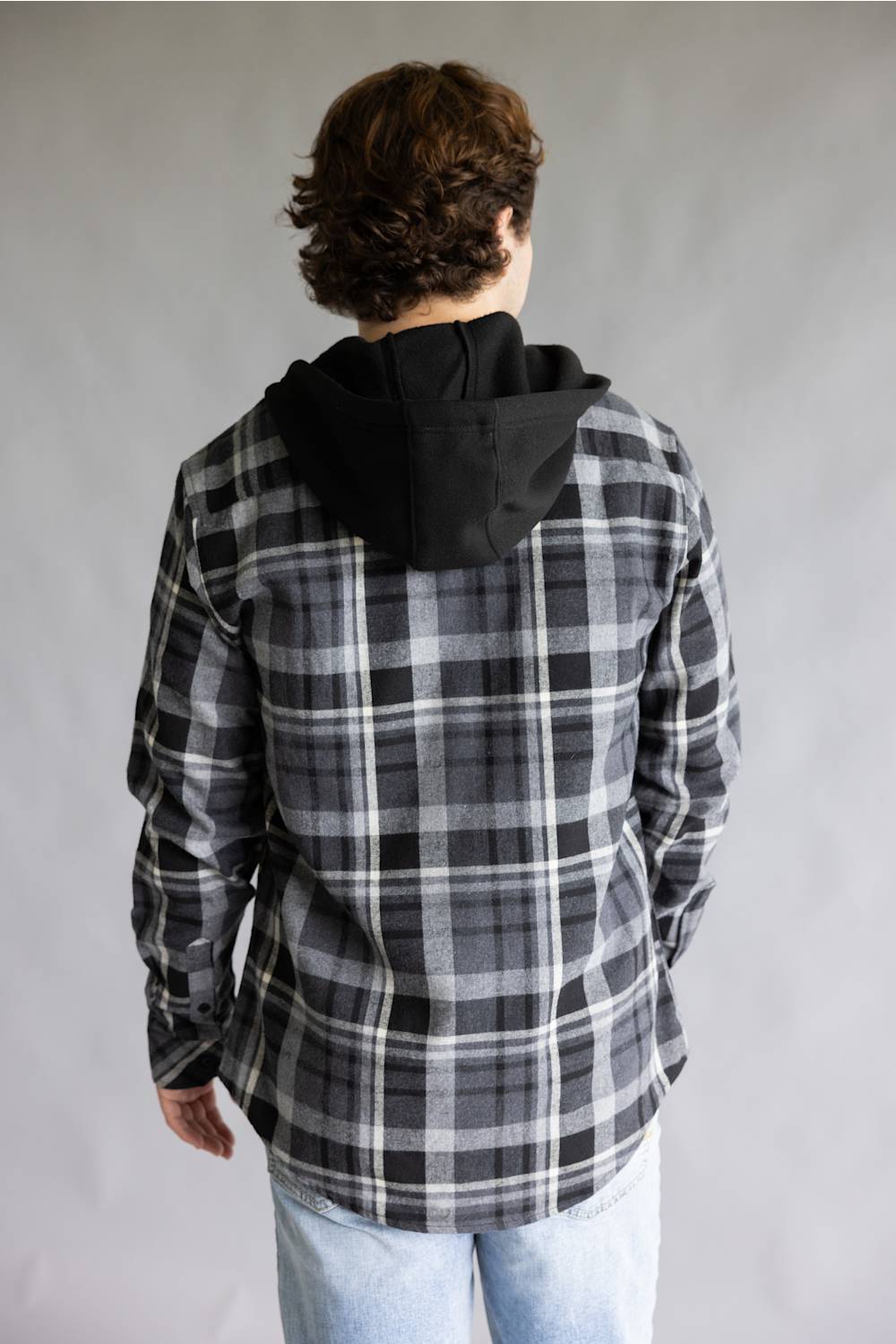 Back view of a model wearing the Split Men's long-sleeve hooded flannel shirt in a black, grey, and white plaid pattern, featuring a solid black attached hood and a straight back yoke against a grey studio background.