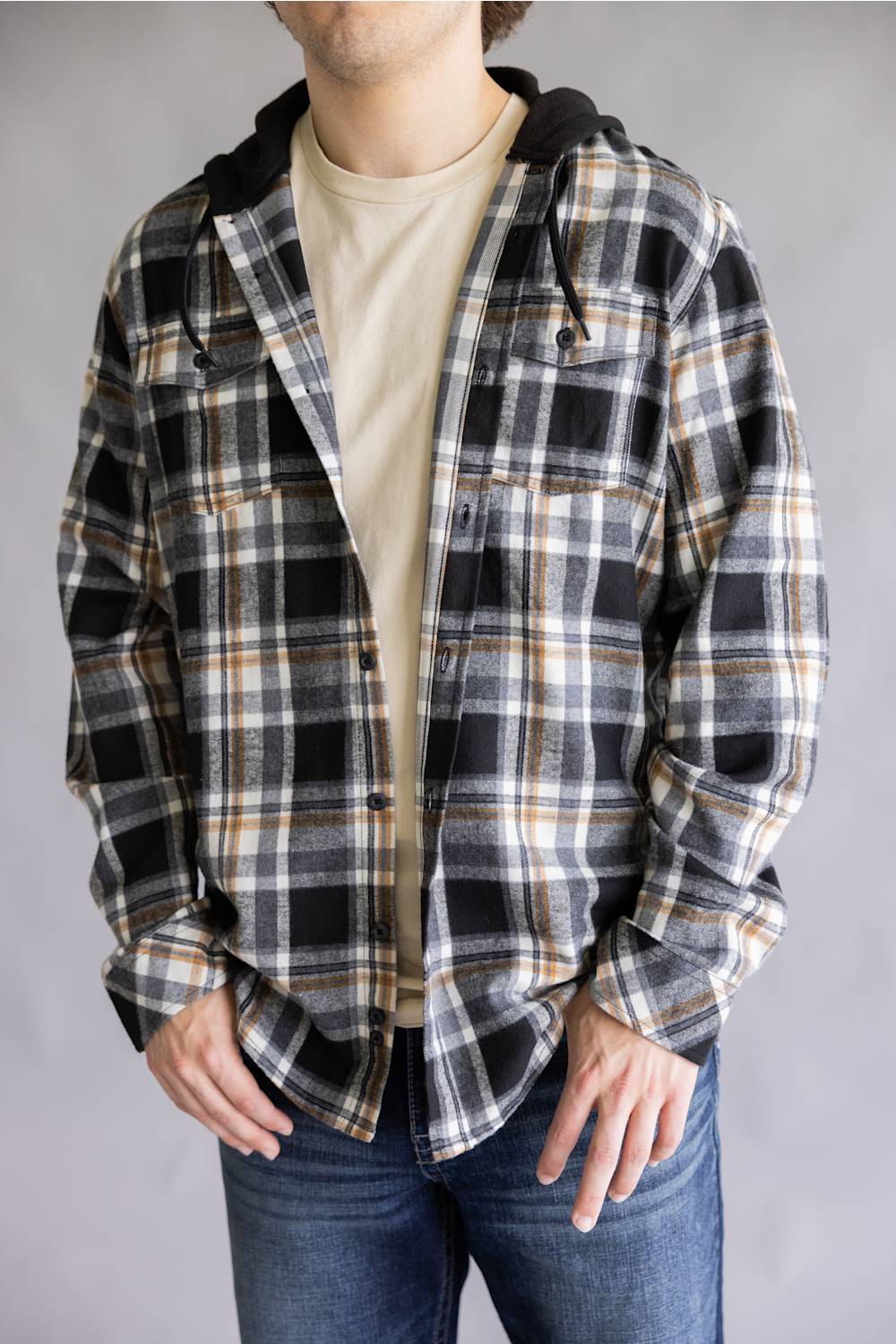 Close-up front view of a model wearing the Split Men's long-sleeve unbuttoned hooded flannel shirt in a cream, black, and tan plaid pattern, styled over an off-white t-shirt with hands in blue jean pockets against a gray studio background.