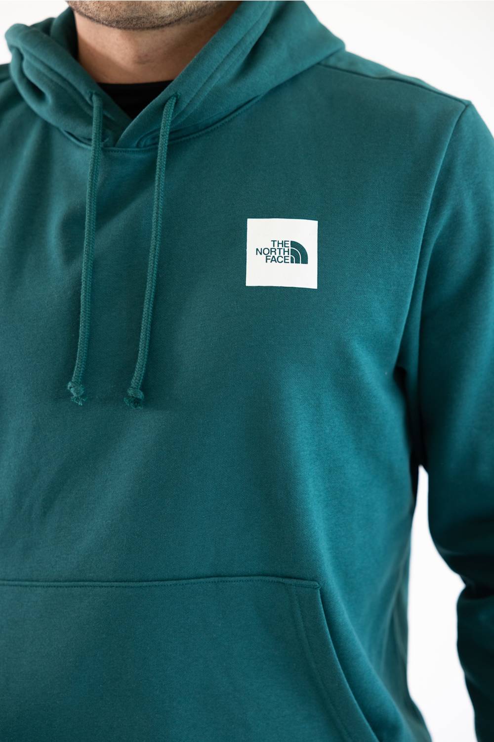 A close-up of a person wearing a teal green pullover hoodie with a white square The North Face logo patch on the left chest.