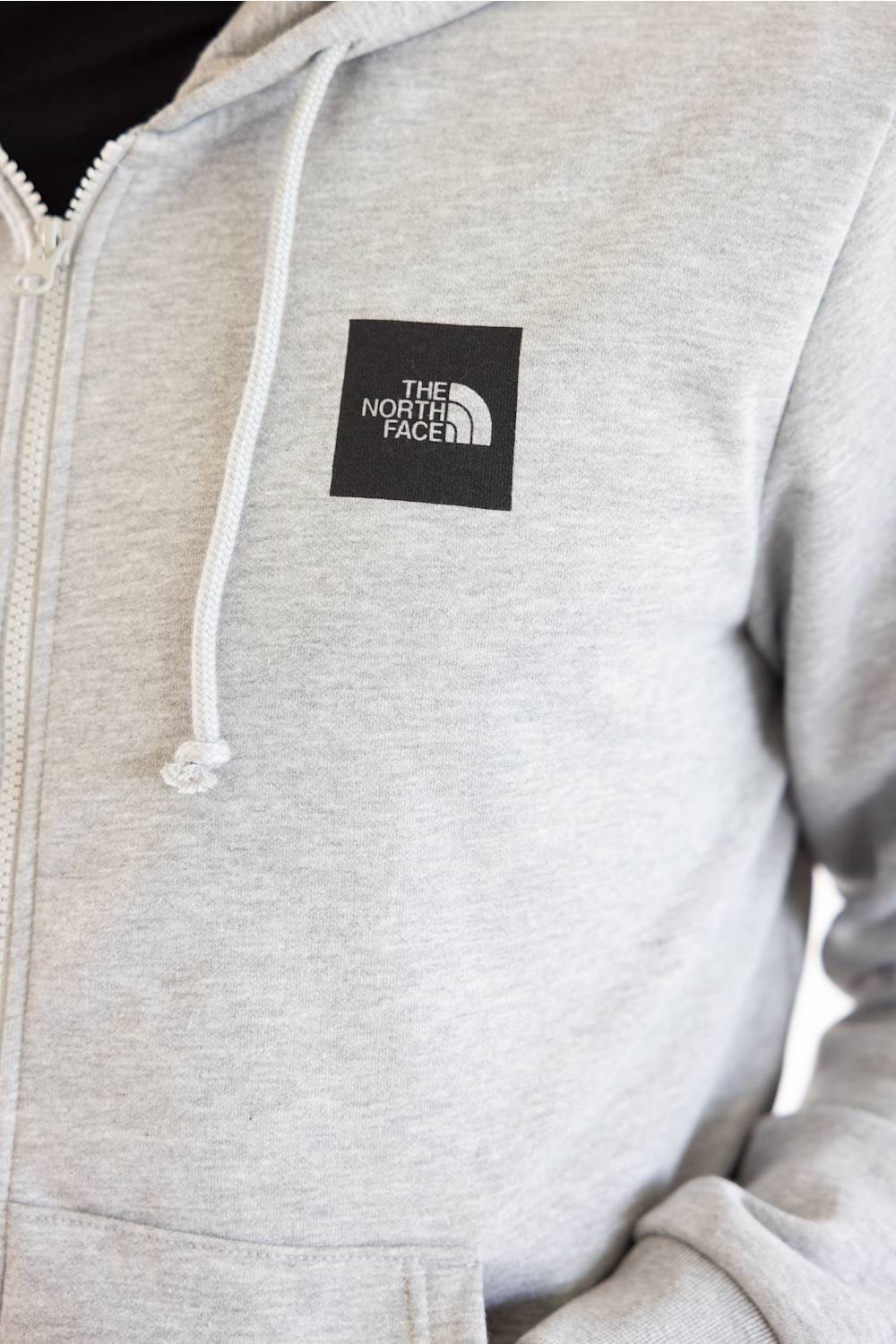 Close-up of a light heather gray zip-up hoodie with white drawstrings and a black square The North Face logo patch on the left chest.