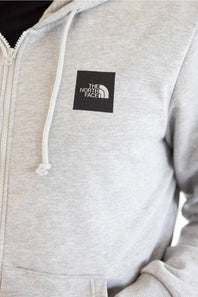 Close-up of a light heather gray zip-up hoodie with white drawstrings and a black square The North Face logo patch on the left chest.