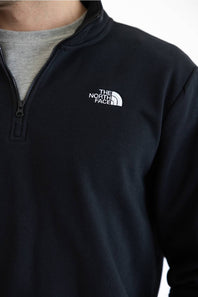 A close up of a man wearing a black long-sleeve quarter-zip pullover jacket and blue jeans, set against a plain white studio background.
