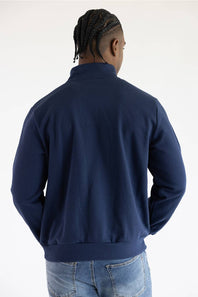 A man viewed from behind wearing a navy blue quarter-zip sweatshirt and blue jeans against a plain white studio background.