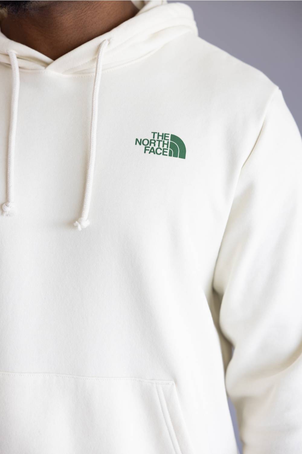 A close-up of a person wearing a cream-colored hoodie with white drawstrings and a green embroidered The North Face logo on the left chest.