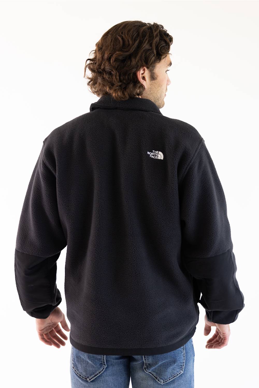 A man viewed from the back wearing a black fleece pullover jacket with a small white The North Face logo on the upper back, paired with blue jeans against a plain white background.