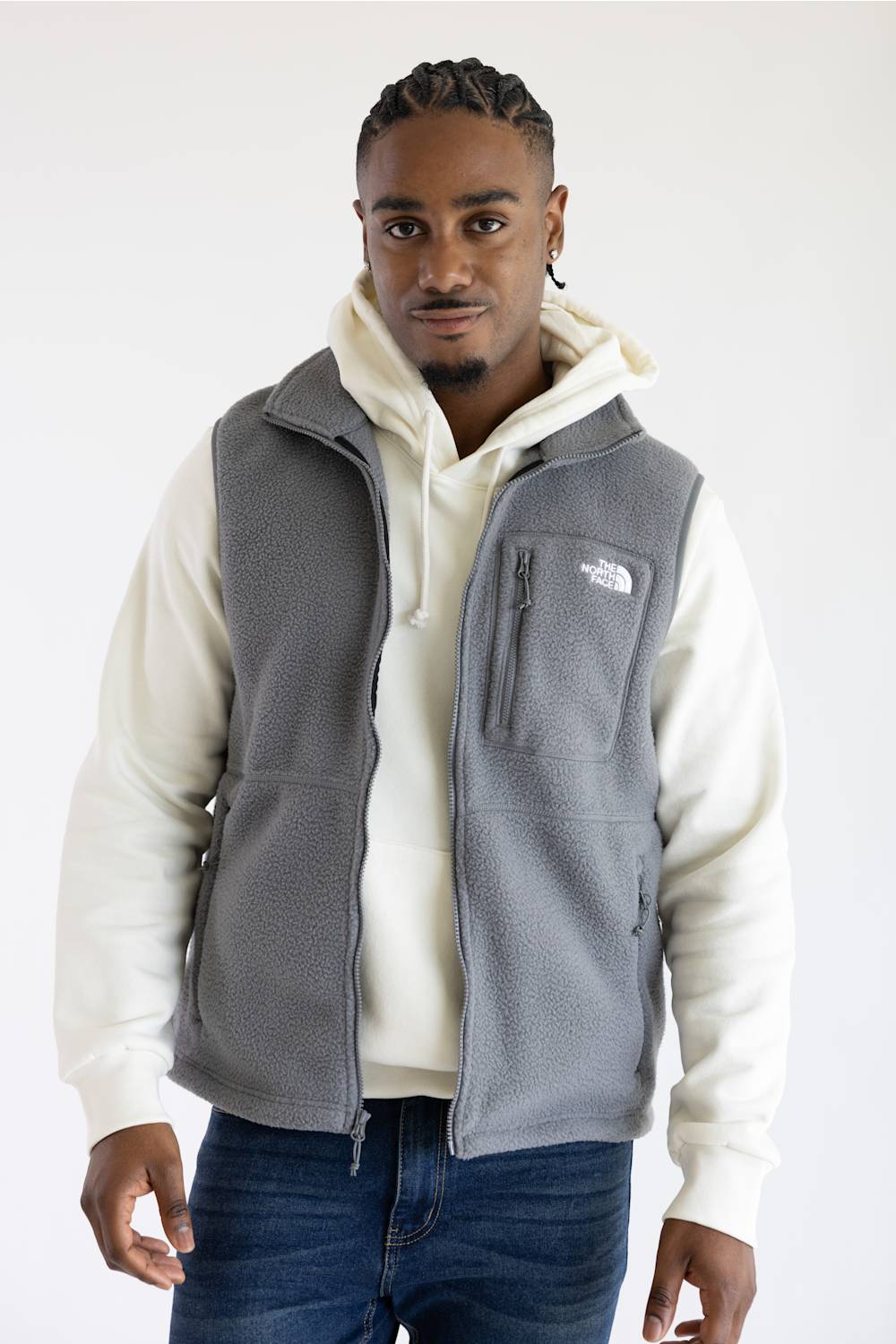 A man wearing a gray fleece vest over a cream zip-up hoodie and blue jeans, standing against a plain white studio background. The vest has a small white embroidered logo on the left chest.