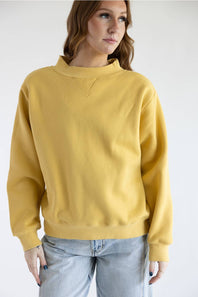A woman wearing a mustard yellow crewneck sweatshirt and light wash jeans, photographed against a plain white studio background.