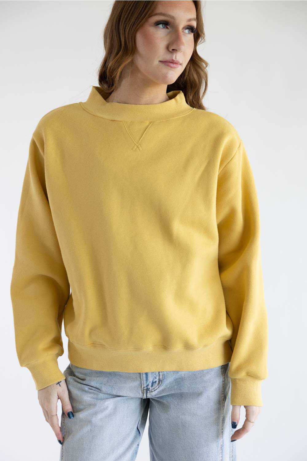 A woman wearing a mustard yellow crewneck sweatshirt and light wash jeans, photographed against a plain white studio background.