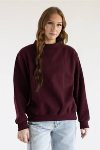 A woman with red hair wearing a loose-fitting, long-sleeve crewneck sweater in deep burgundy and blue jeans, standing against a plain white background.
