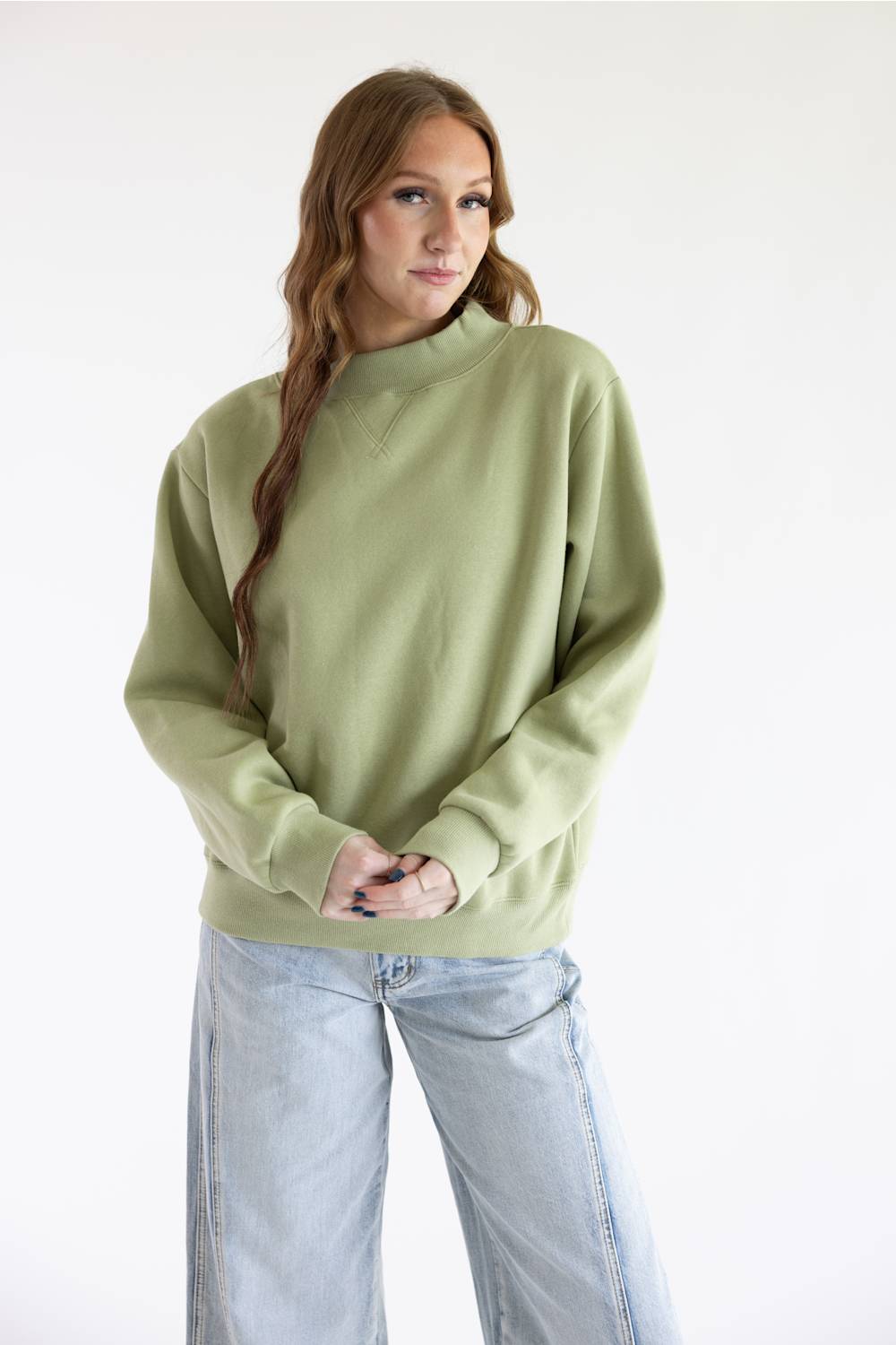 A woman modeling a sage green mock neck sweatshirt with light-wash wide-leg jeans against a plain white studio background.