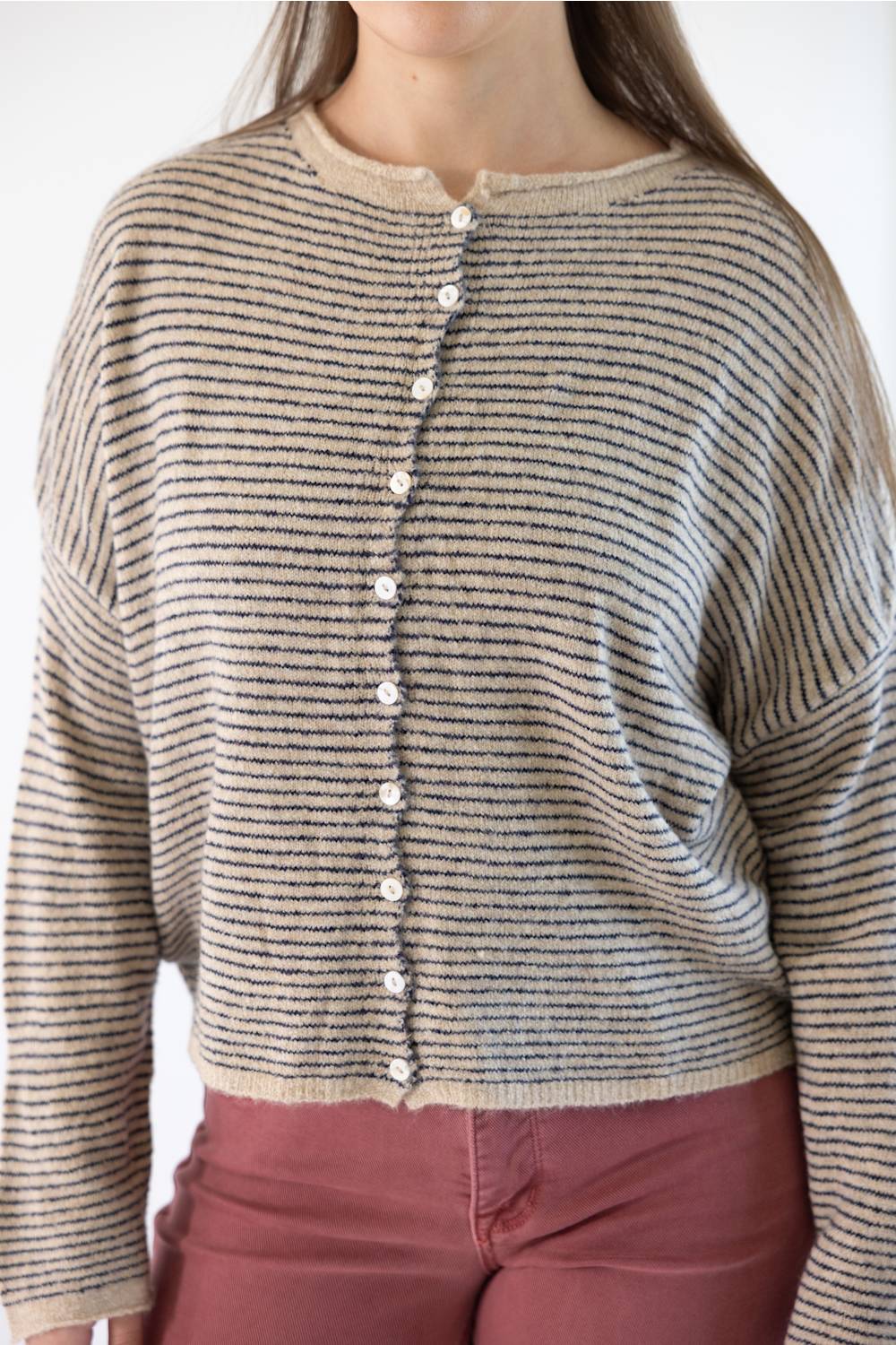 A person wearing a cropped knit beige cardigan with thin horizontal stripes, featuring a button-down front. The cardigan is paired with dusty rose pants against a plain white background.