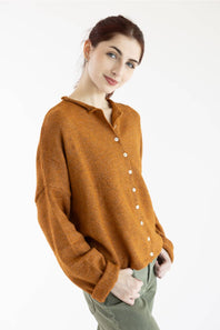 A woman wearing a long-sleeve knit cardigan in a warm rust-orange color with white buttons, paired with olive green pants, against a plain white background.