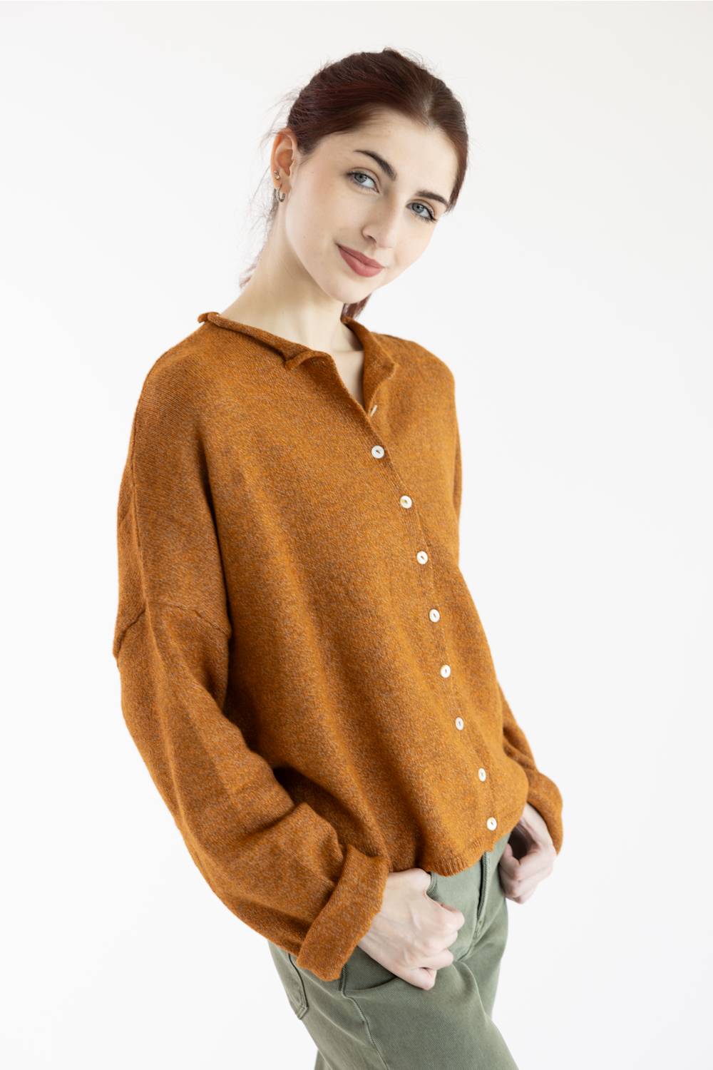 A woman wearing a long-sleeve knit cardigan in a warm rust-orange color with white buttons, paired with olive green pants, against a plain white background.