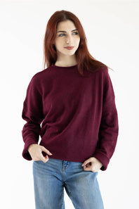 A woman with red hair wearing a maroon crewneck knit sweater and blue jeans, standing against a plain white background.