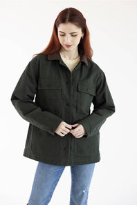 A woman with red hair wearing an oversized dark olive green utility jacket with a brown collar, styled with a cream ribbed sweater and blue jeans, against a plain white background.
