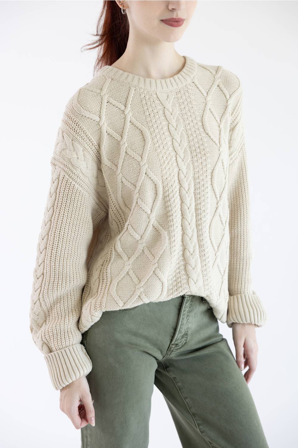 A woman wearing a cream-colored cable-knit sweater and olive green pants, shown from the neck down against a plain white background.