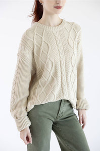 A woman wearing a cream-colored cable-knit sweater and olive green pants, shown from the neck down against a plain white background.