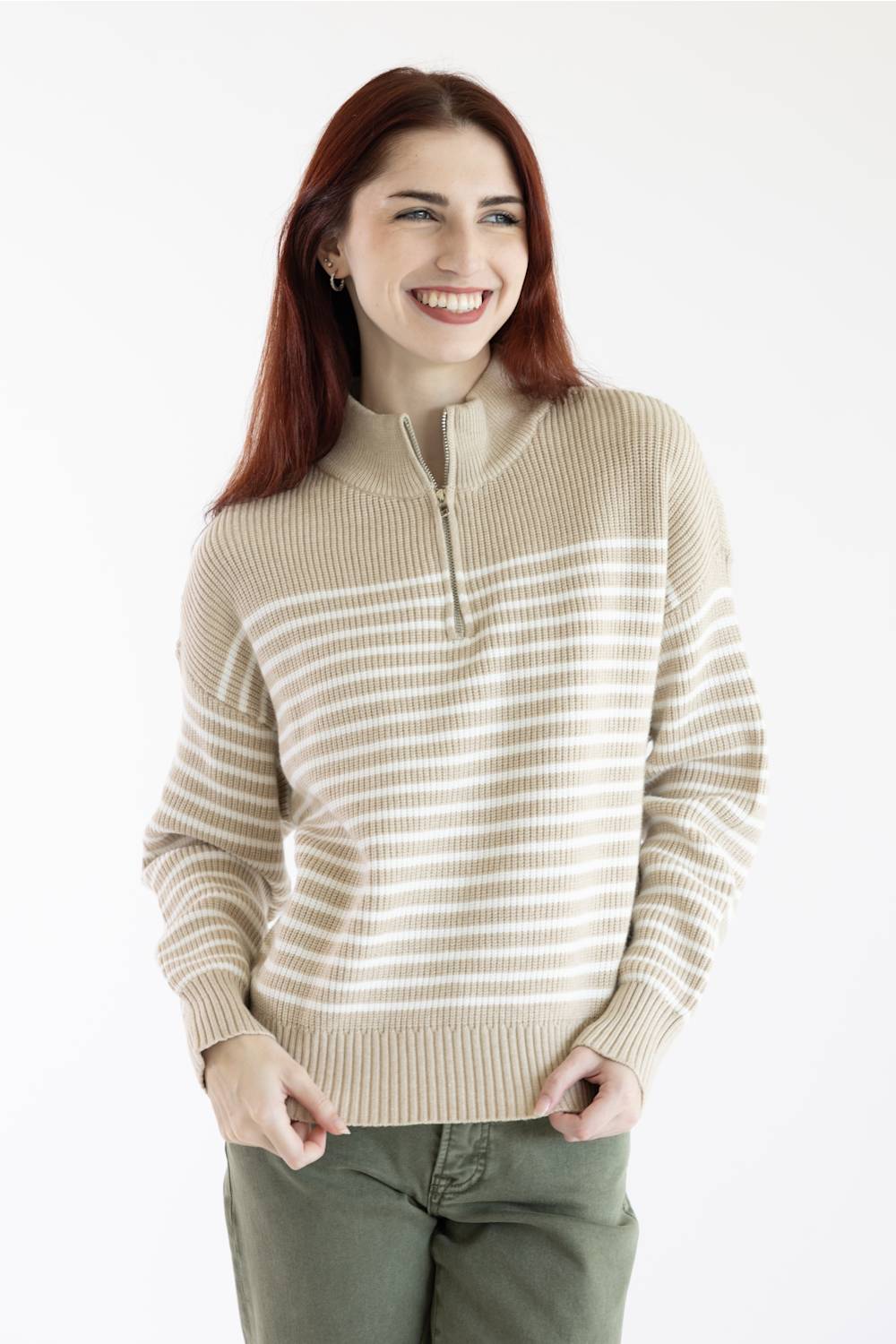 A smiling woman with red hair wearing a beige and white horizontally striped quarter-zip sweater and olive green pants, photographed against a plain white studio background.